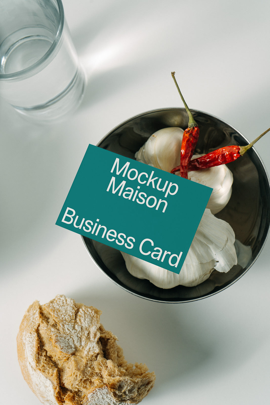 PSD mockup of a business card placed on top of garlic and red chili peppers in a bowl, next to a glass of water and a piece of bread.