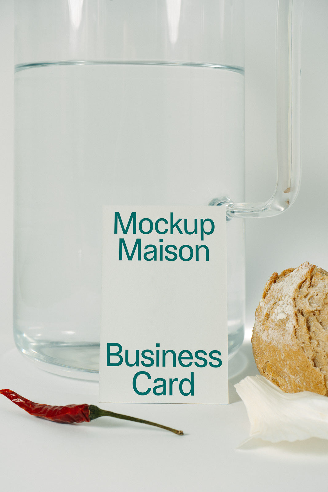 PSD mockup of a business card standing against a glass pitcher with water, next to a round loaf of bread and a red chili on a white surface.
