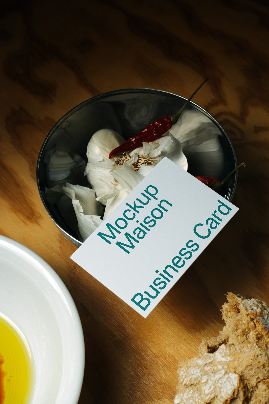 PSD mockup of a business card with "Mockup Maison" text, placed on a wooden surface near a bowl of garlic with chili and a piece of bread.