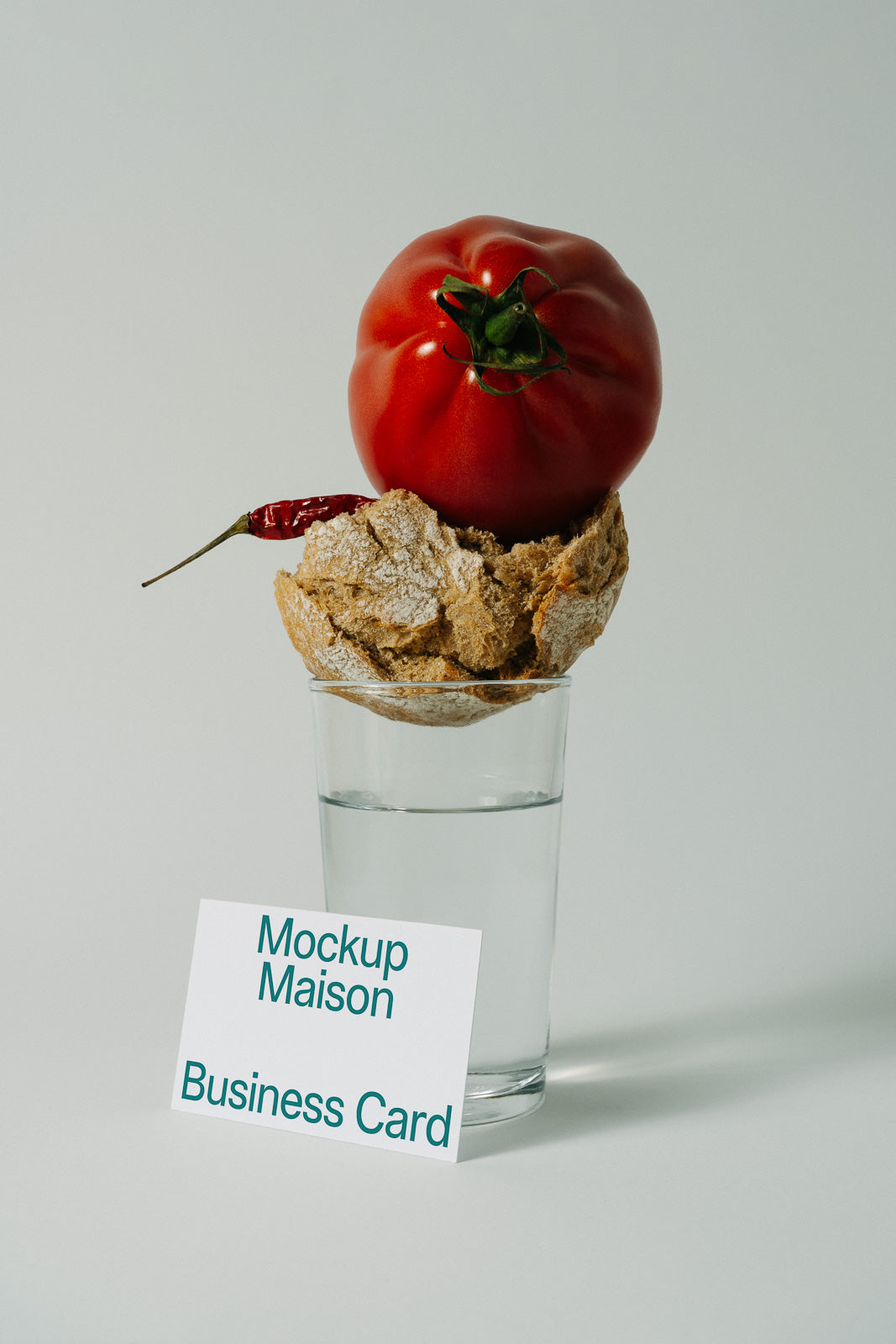 PSD mockup of a business card leaning against a glass filled with water, topped by a round loaf of bread, a red tomato, and a chili pepper.