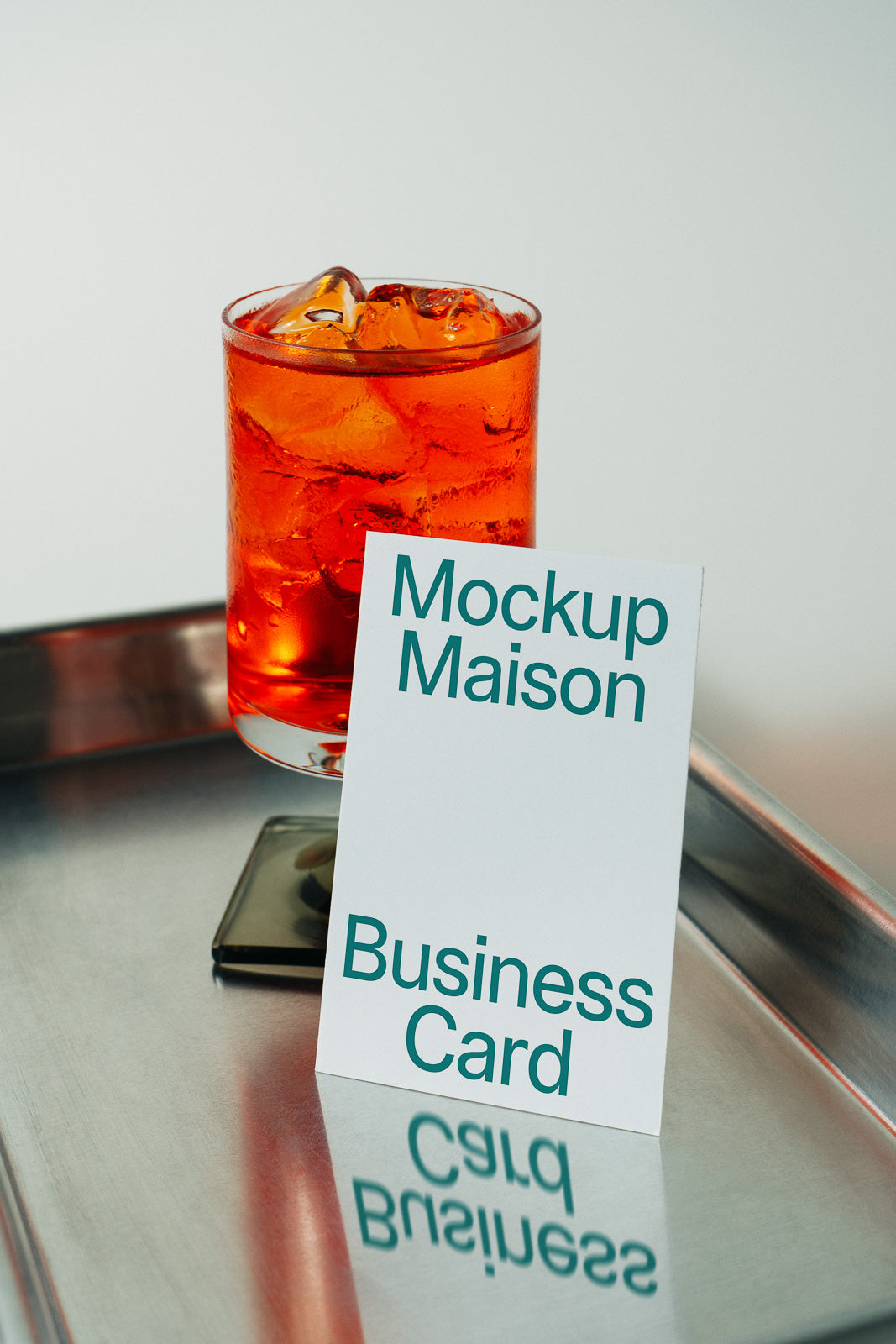 PSD mockup of a business card standing on a metal tray beside a glass filled with an orange beverage and ice cubes, reflecting the card's text.