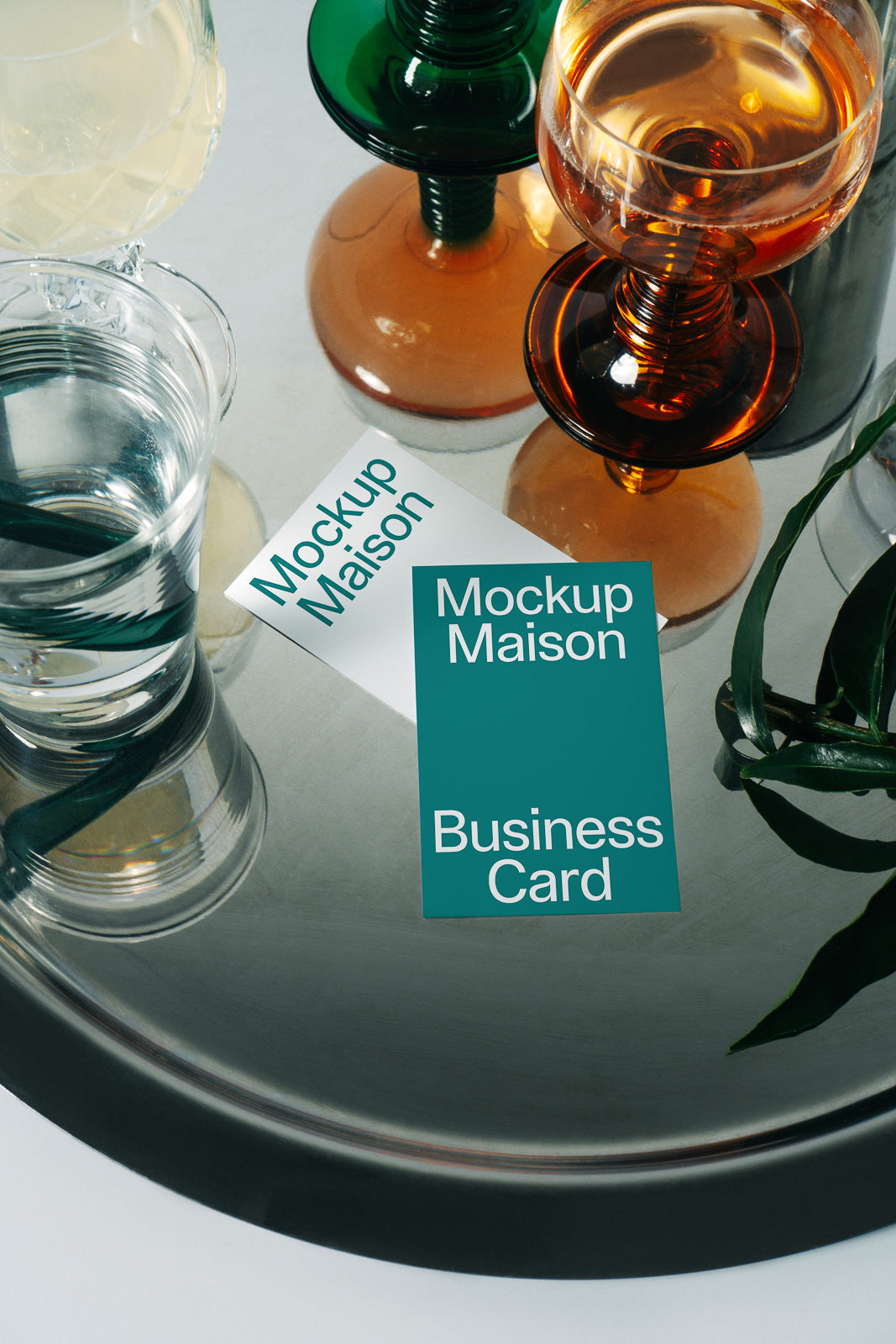 PSD mockup of business cards labeled "Mockup Maison" on a reflective tray, surrounded by colorful glassware and a green plant leaf.
