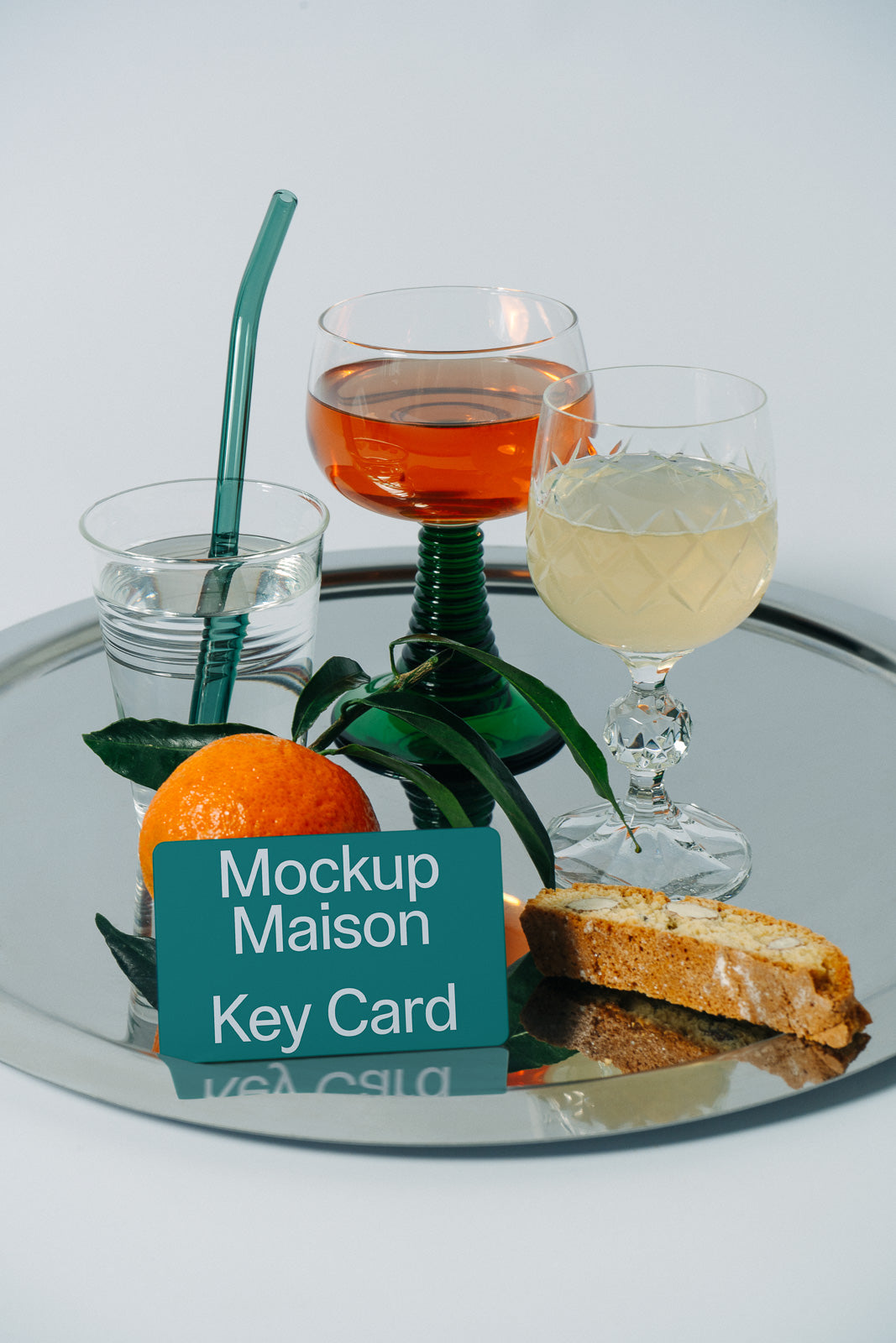 PSD mockup of a key card on a mirrored tray with a glass of water, orange and yellow drinks, an orange fruit, and biscotti.