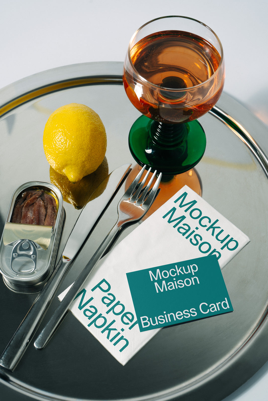 PSD mockup of a branded paper napkin and business card alongside a fork, knife, tinned anchovies, lemon, and a drink on a silver tray.