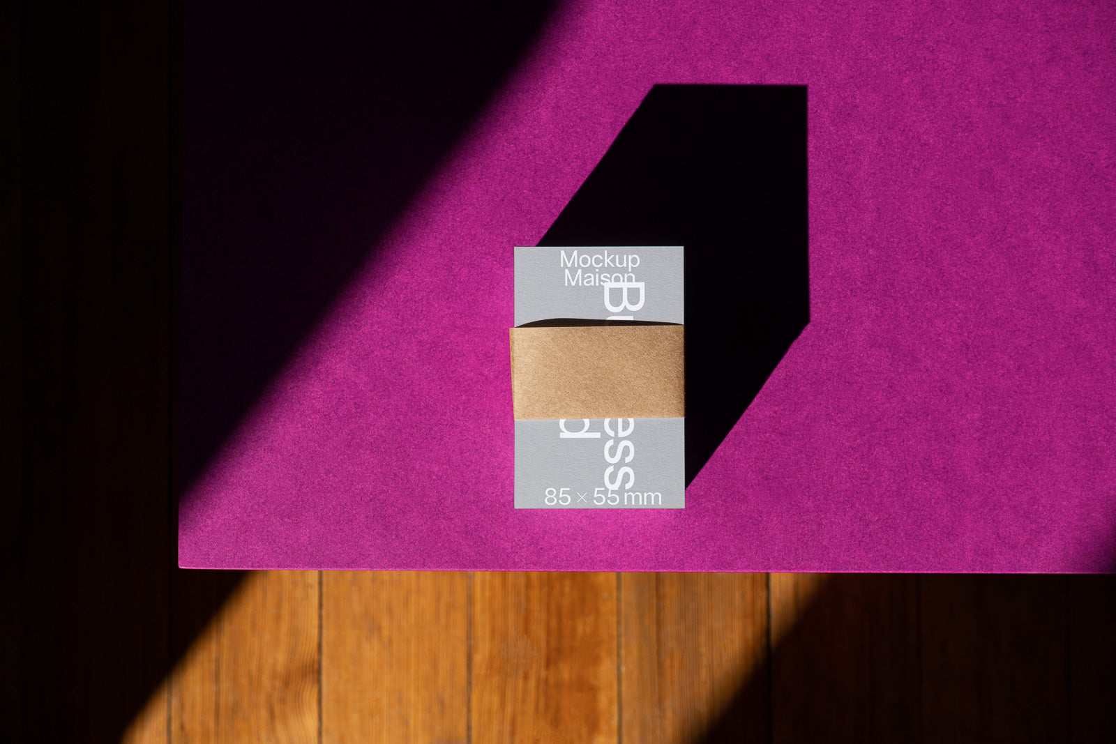 PSD mockup of a business card wrapped with a brown paper band on a purple surface, casting a shadow on a wooden floor.