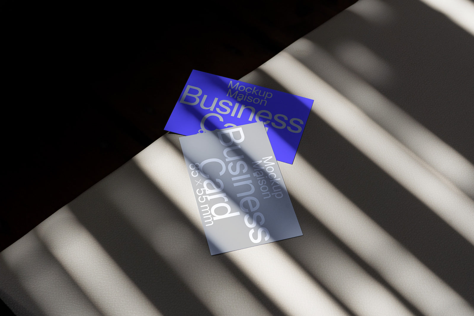 PSD mockup of two business cards on a textured surface, one blue and one transparent, with light and shadow patterns cast across them.