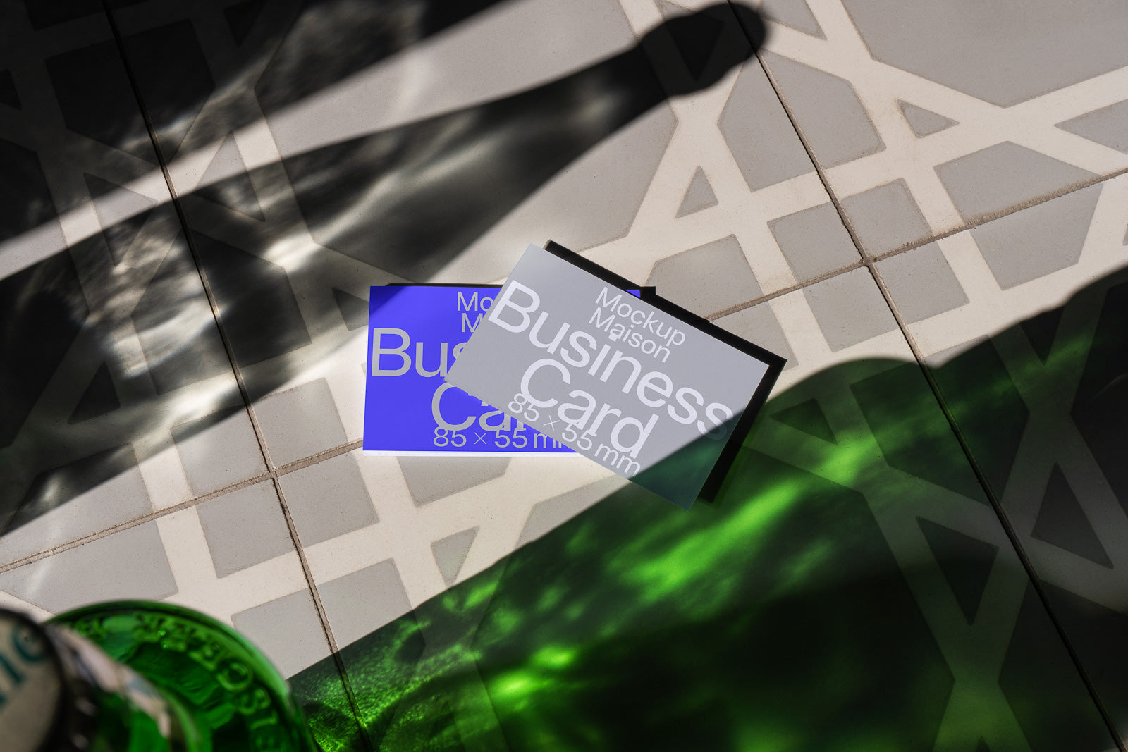 PSD mockup of two business cards in 85x55 mm size, lying on a geometric patterned surface with sunlight and green shadows creating a dynamic effect.