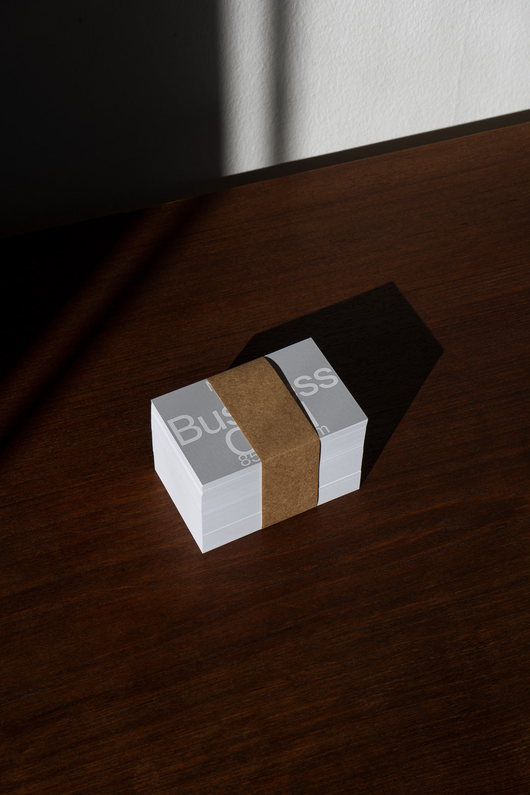 PSD mockup of a stack of business cards wrapped with a brown paper band, placed on a wooden surface with dramatic lighting creating shadows.