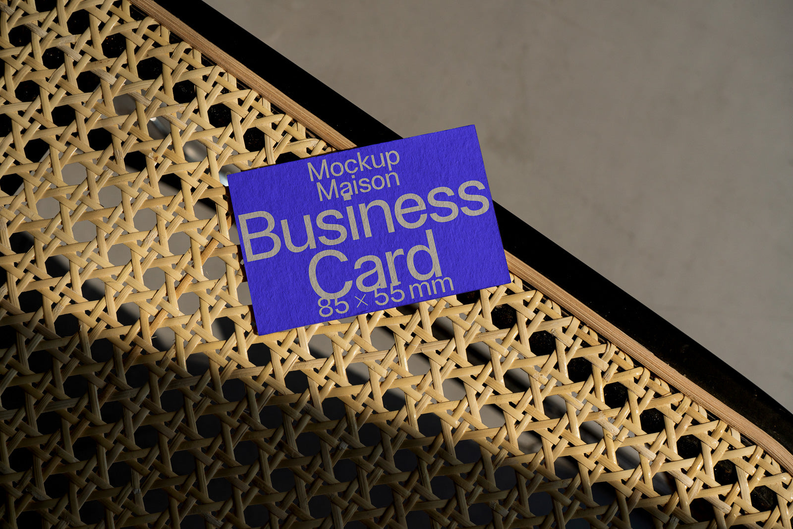 PSD mockup of a blue business card measuring 85 × 55 mm, resting on a woven chair surface with intricate patterns, casting shadows.