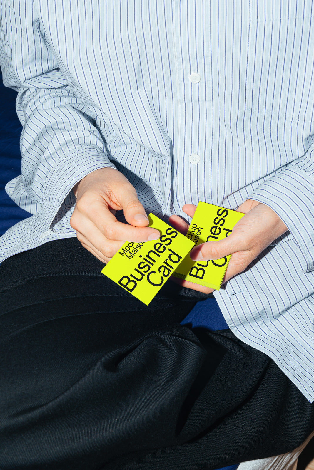 PSD mockup of a person holding two bright yellow business cards, dressed in a striped shirt and black pants, sitting on a dark surface.