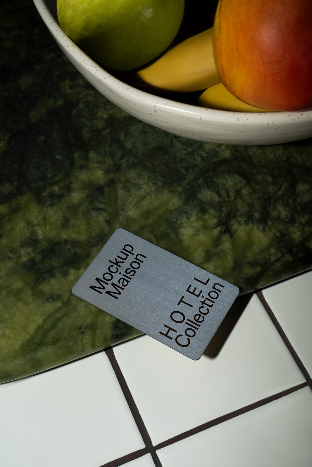 PSD mockup of a card labeled "Mockup Maison HOTEL Collection" lying on a dark green marble surface beside a ceramic bowl of fruit, including bananas and apples.
