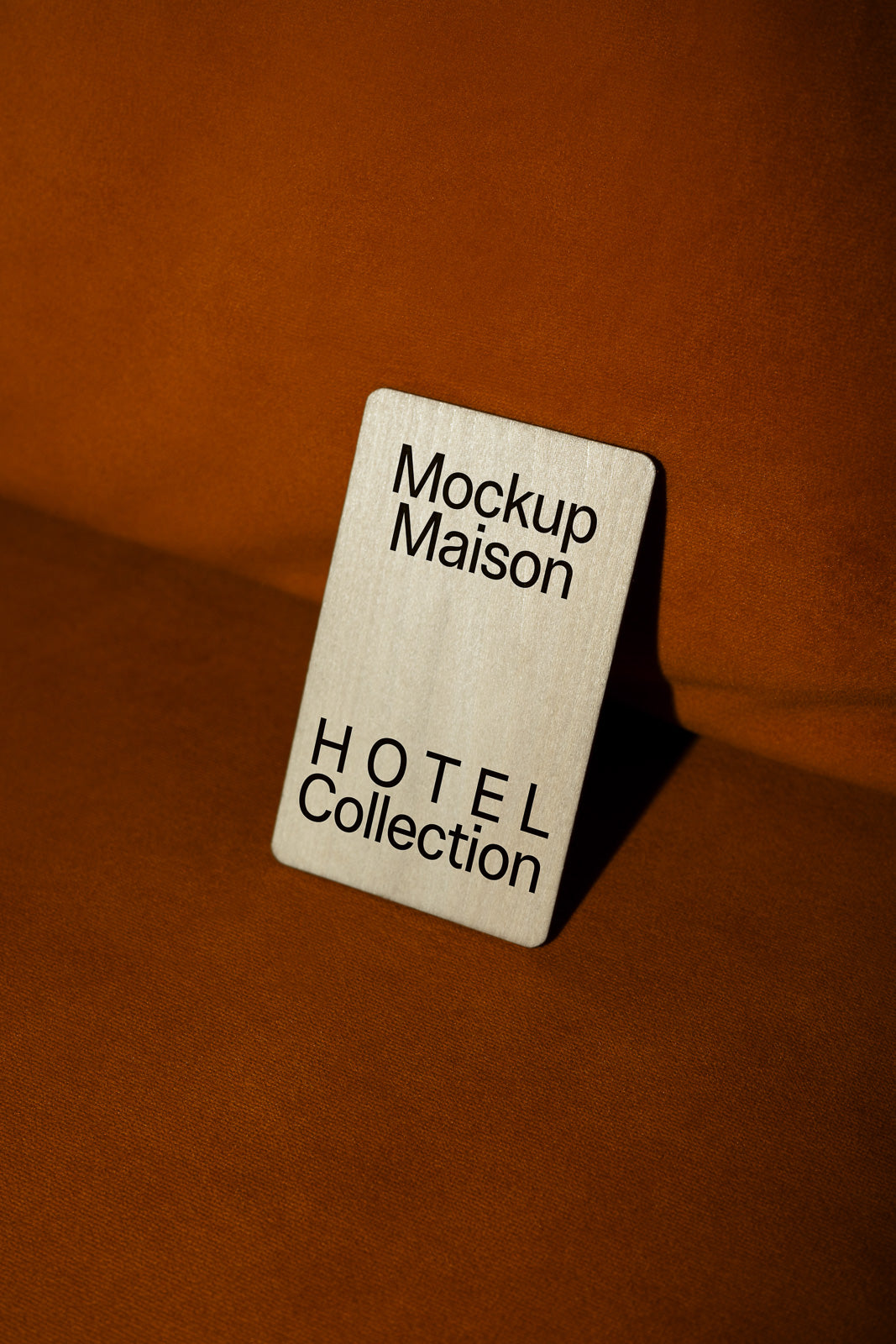 PSD mockup of a wooden card displaying the text "Mockup Maison Hotel Collection" on a brown fabric background.