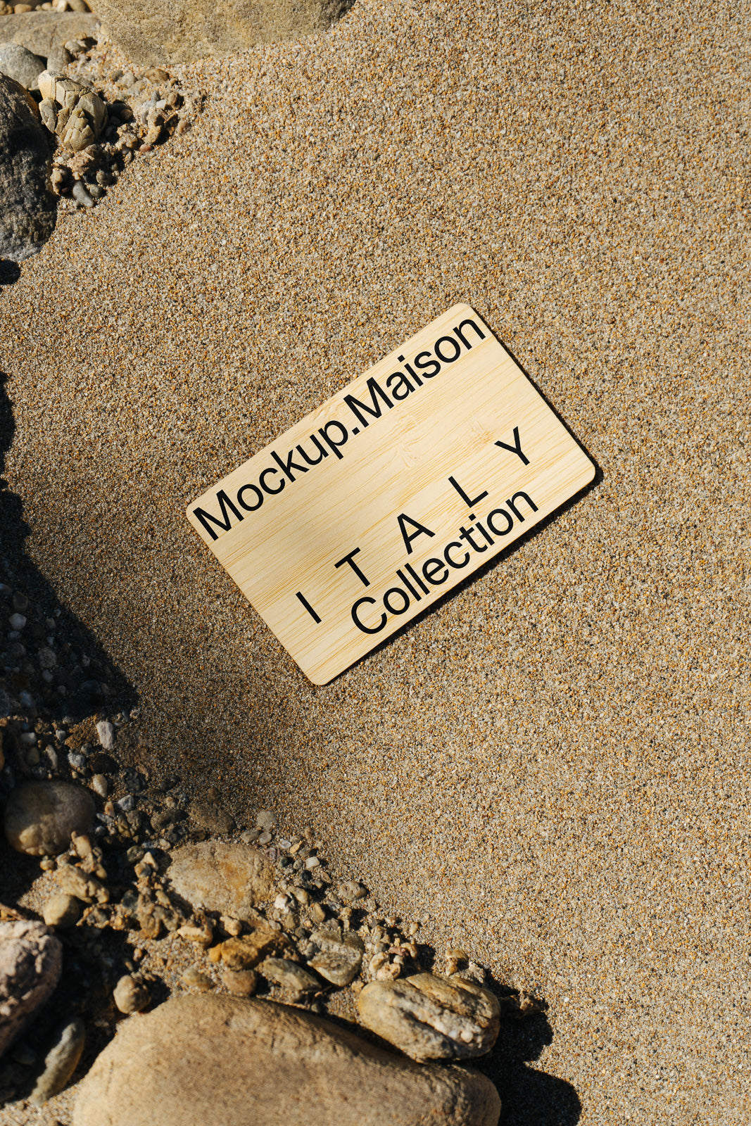PSD mockup of a wooden sign labeled "Mockup.Maison ITALY Collection" placed on textured sandy beach with scattered stones.