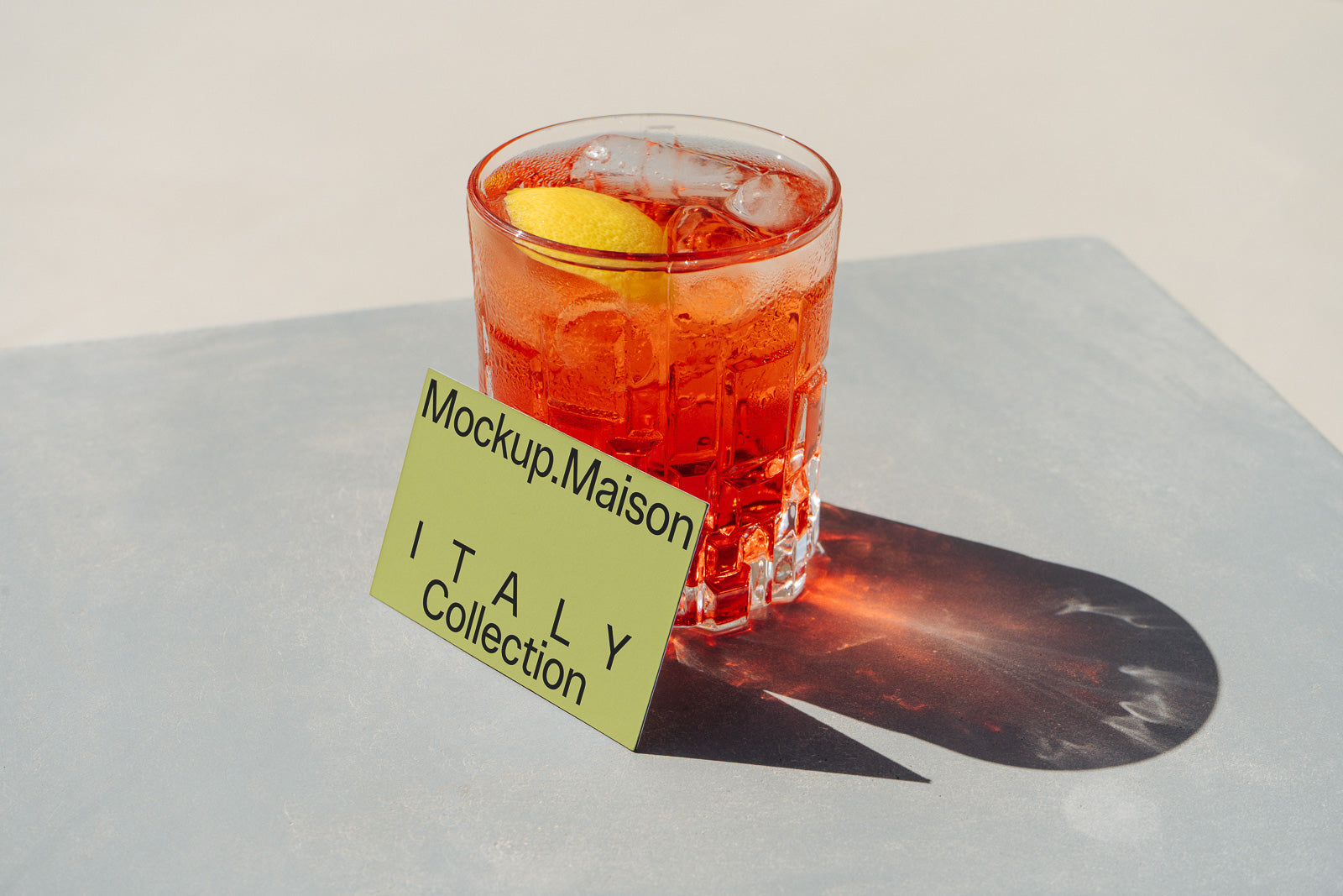 PSD mockup of a glass filled with an orange-colored drink and ice, garnished with a lemon slice. Next to the glass is a card labeled "Mockup.Maison ITALY Collection."