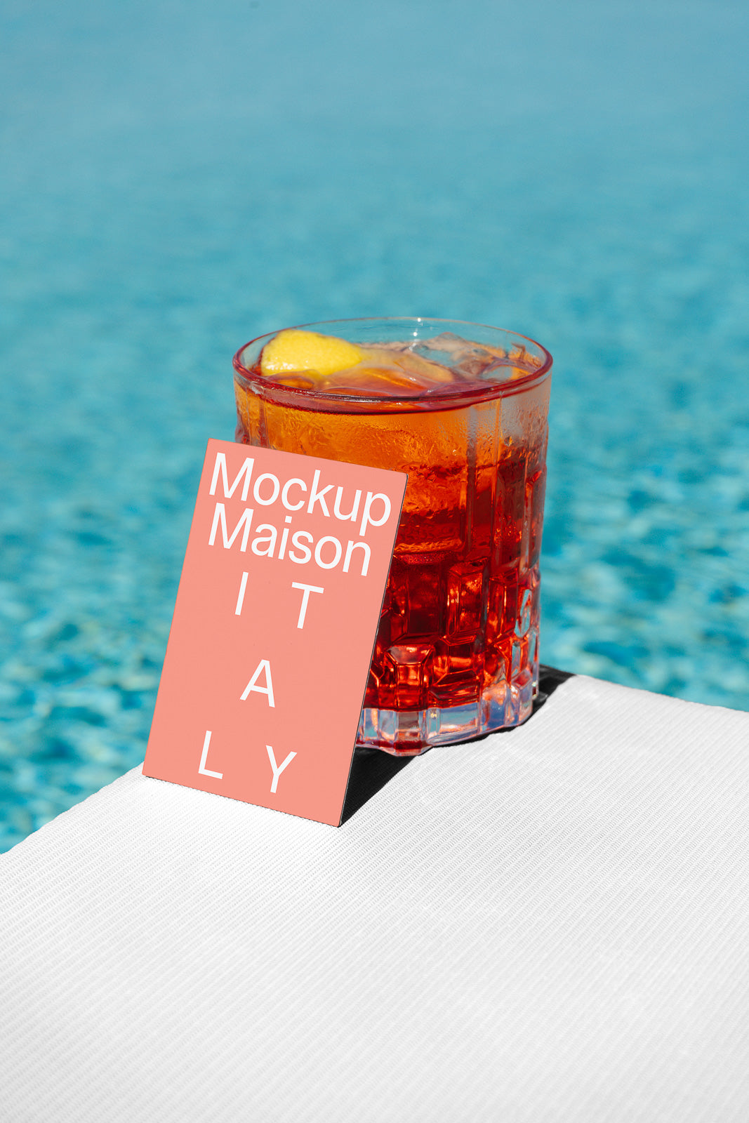 PSD mockup of a card labeled "Mockup Maison Italy" placed next to a glass with a red drink and lemon slice, set against a pool background.