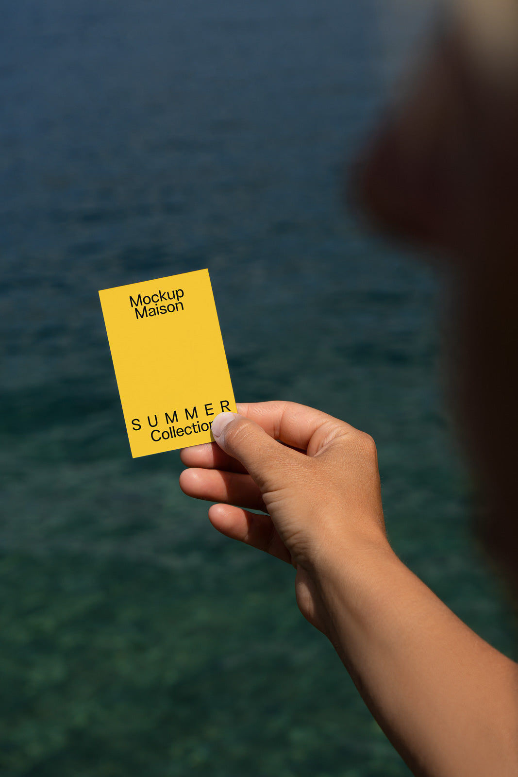 PSD mockup of a hand holding a yellow card with the text "Mockup Maison SUMMER Collection" against a blurry water background.