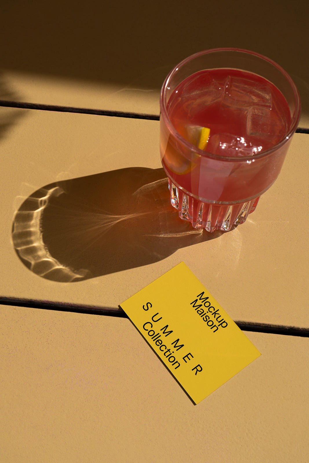 PSD mockup of a pink drink in a glass with ice and a lemon slice on a sunlit table, casting a shadow on a vibrant yellow card labeled "Summer Collection."