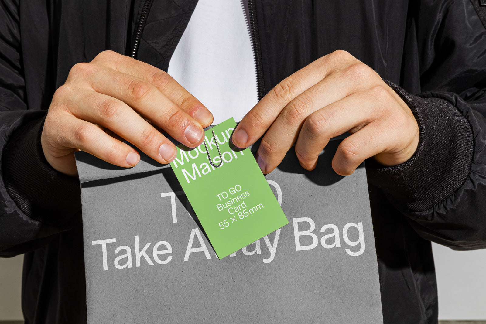 PSD mockup of a person holding a gray takeaway bag with a clipped green business card reading "Mockup Maison TO GO Business Card 55 x 85 mm."