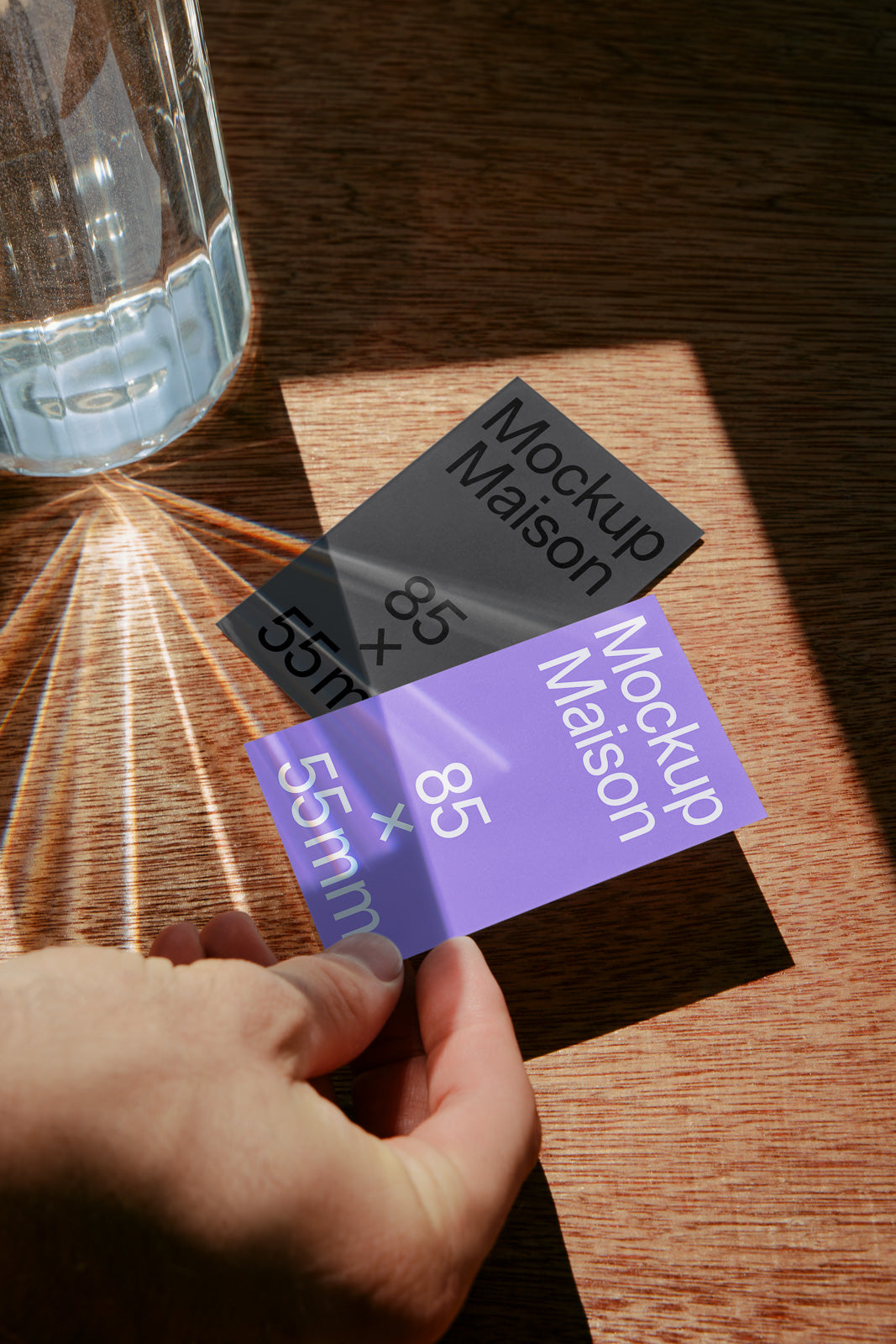 PSD mockup of two business cards on a wooden table, with sunlight casting reflections from a glass, displaying dimensions 85 x 55mm.