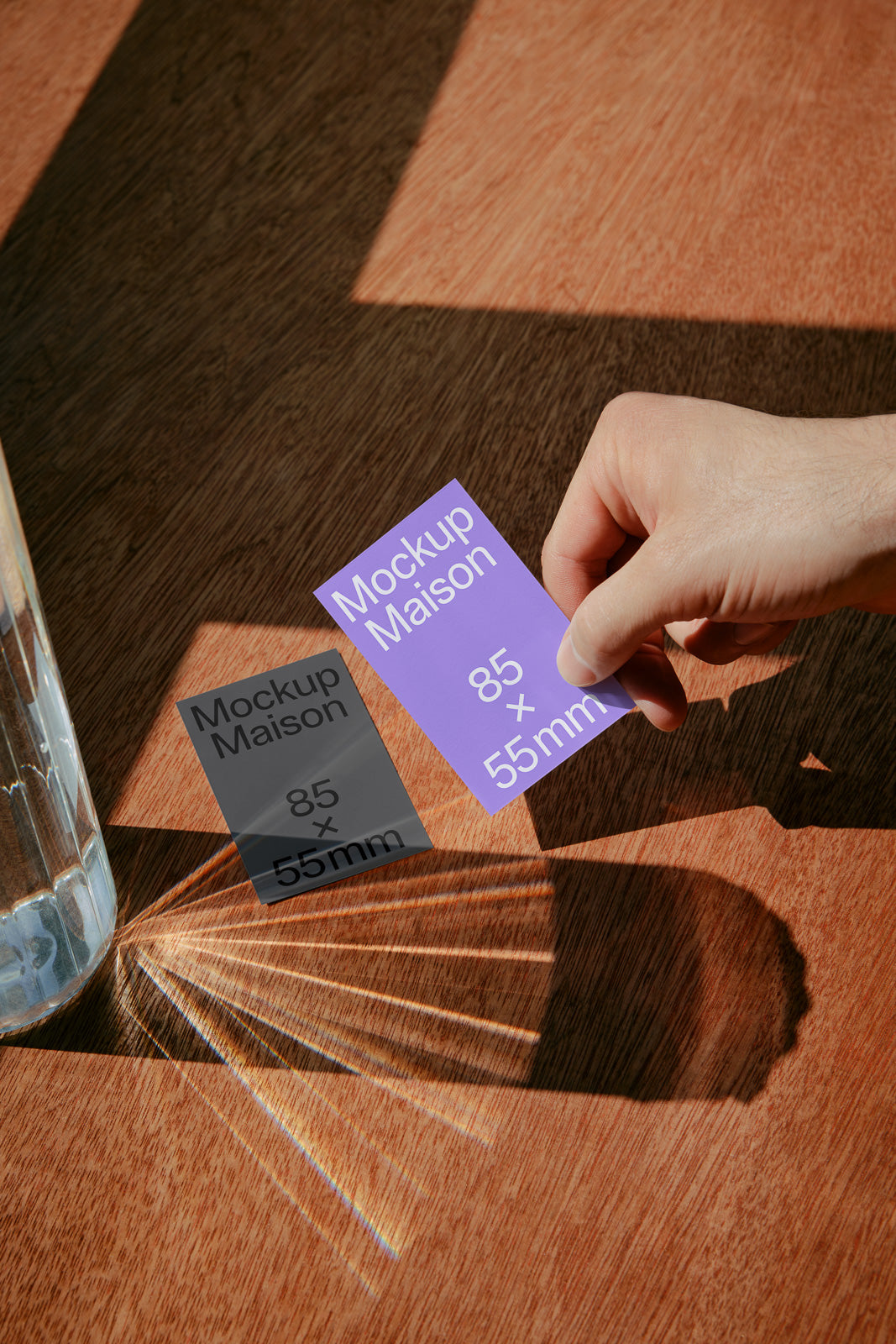 PSD mockup of a hand holding a purple business card with 85 x 55mm text, casting a shadow next to a glass on a wooden table.