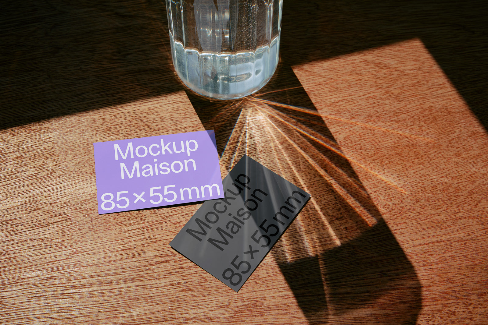 PSD mockup of two business cards on a wooden surface, one purple and one gray, with a glass of water casting dynamic shadows and light reflections.