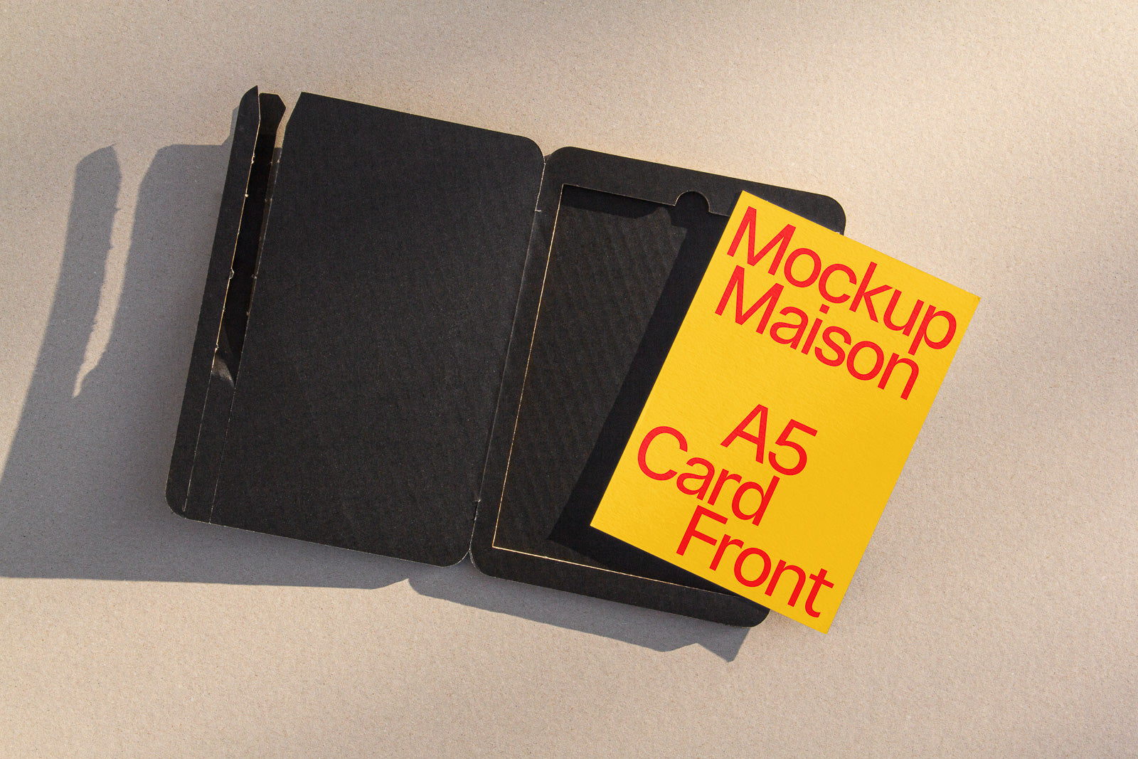 PSD mockup of an A5 card with bold red text on a yellow background, partially inside a black cardboard envelope on a textured surface.