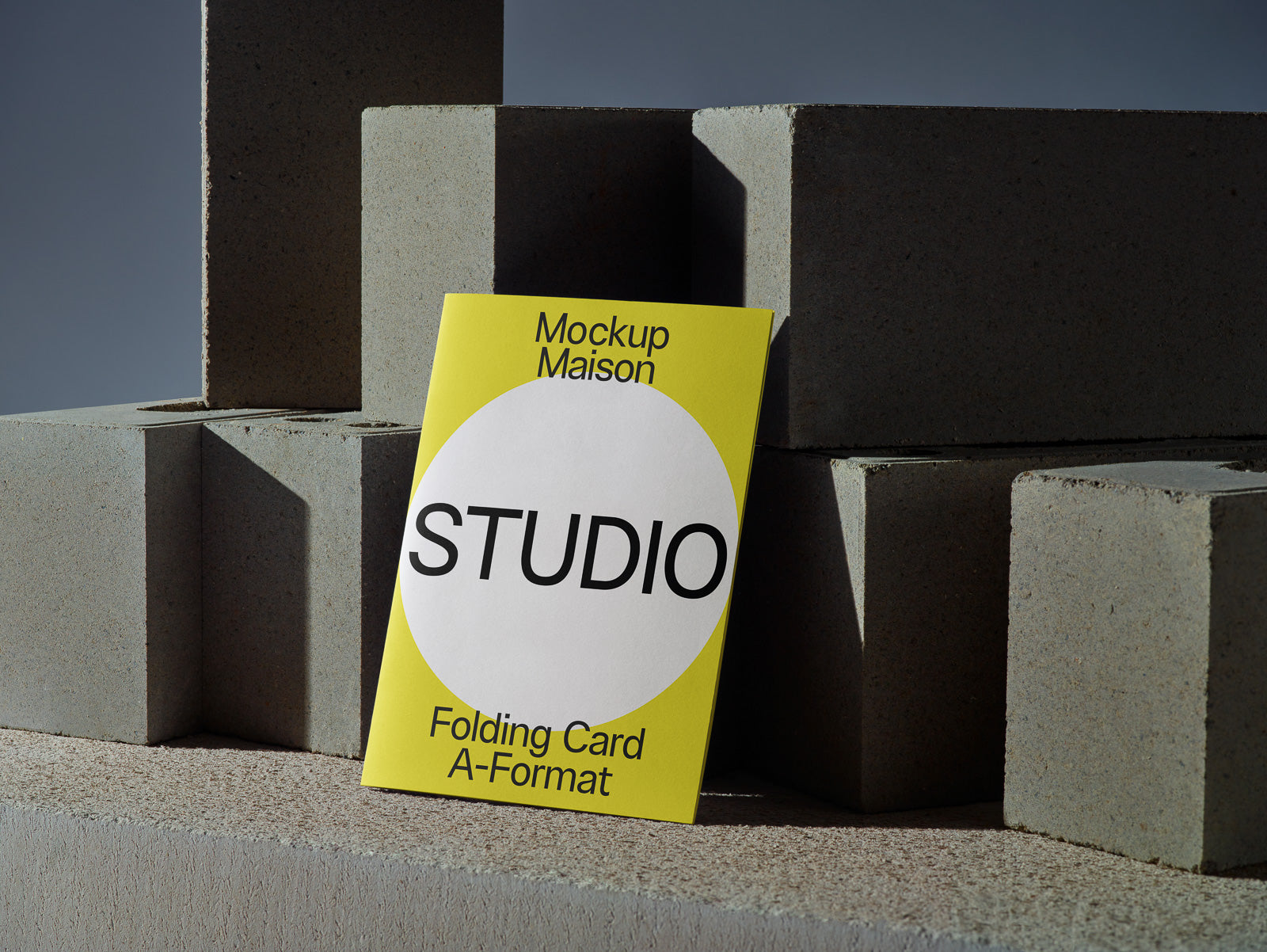 PSD mockup of a folding card in A-format with "Mockup Maison Studio" text in black on a yellow background, leaning against concrete blocks.