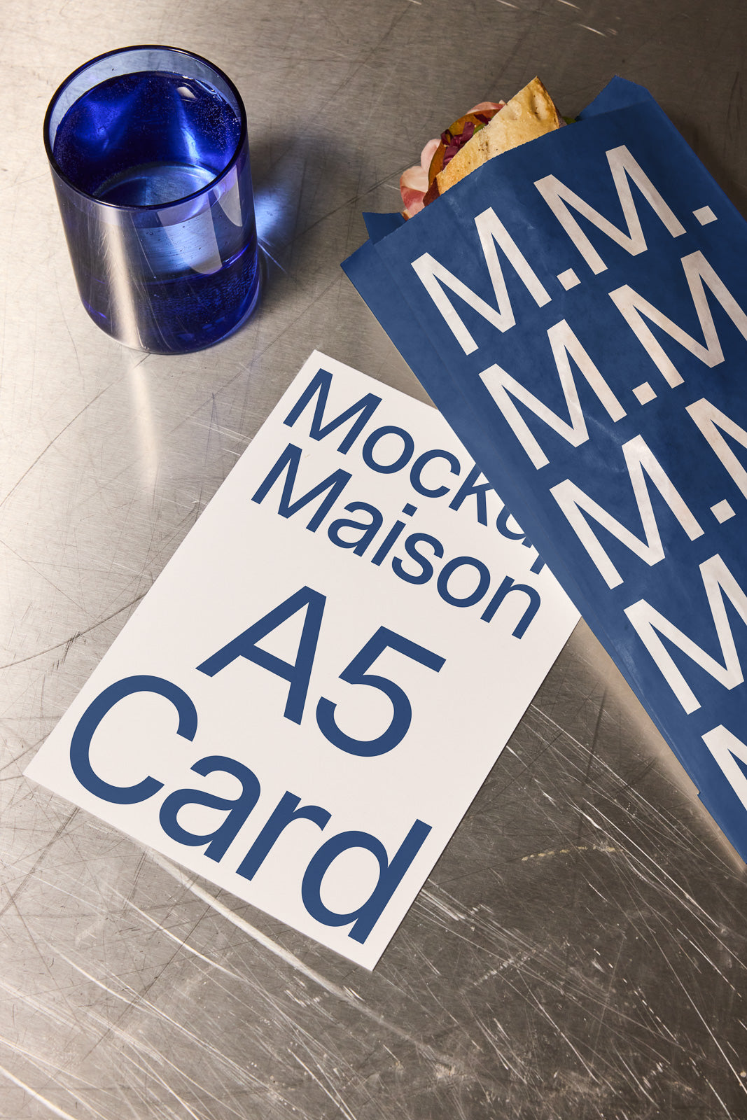 PSD mockup of an A5 card on a metallic surface with a blue glass and sandwich in a blue branded bag.