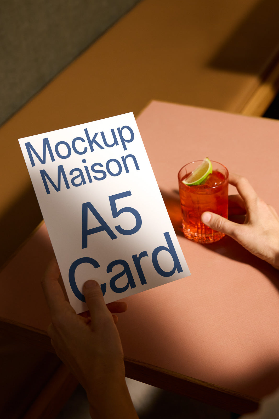 PSD mockup of an A5 card held by a person, with the text "Mockup Maison A5 Card" visible, near a glass containing a drink with a lime wedge.