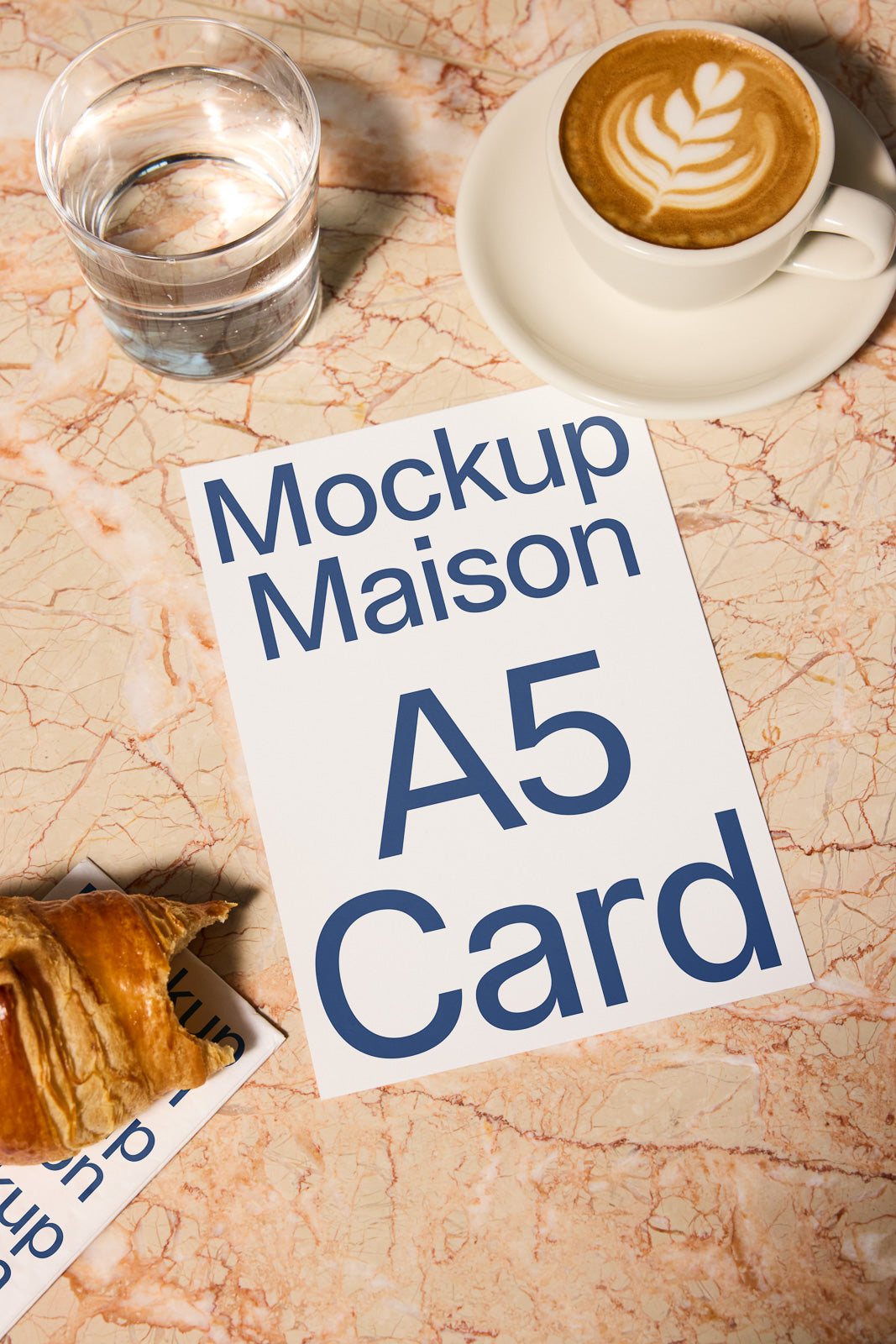 PSD mockup of an A5 card on a marble table near a cappuccino and a glass of water, with a croissant partially covering a card.