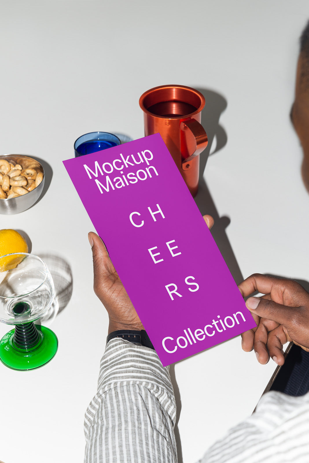 PSD mockup of a person holding a purple card marked "Mockup Maison Cheers Collection" surrounded by colorful glassware and a bowl of cashews on a white table.