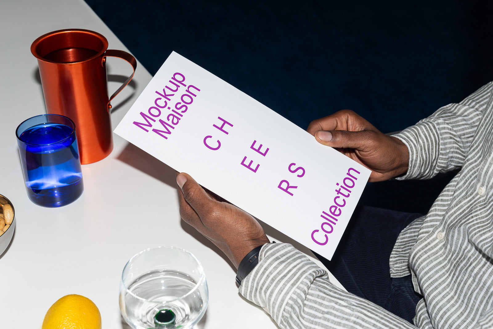 PSD mockup of a person holding a white card with the text "Mockup Maison Cheers Collection." The scene includes a copper jug, blue cup, and lemon.