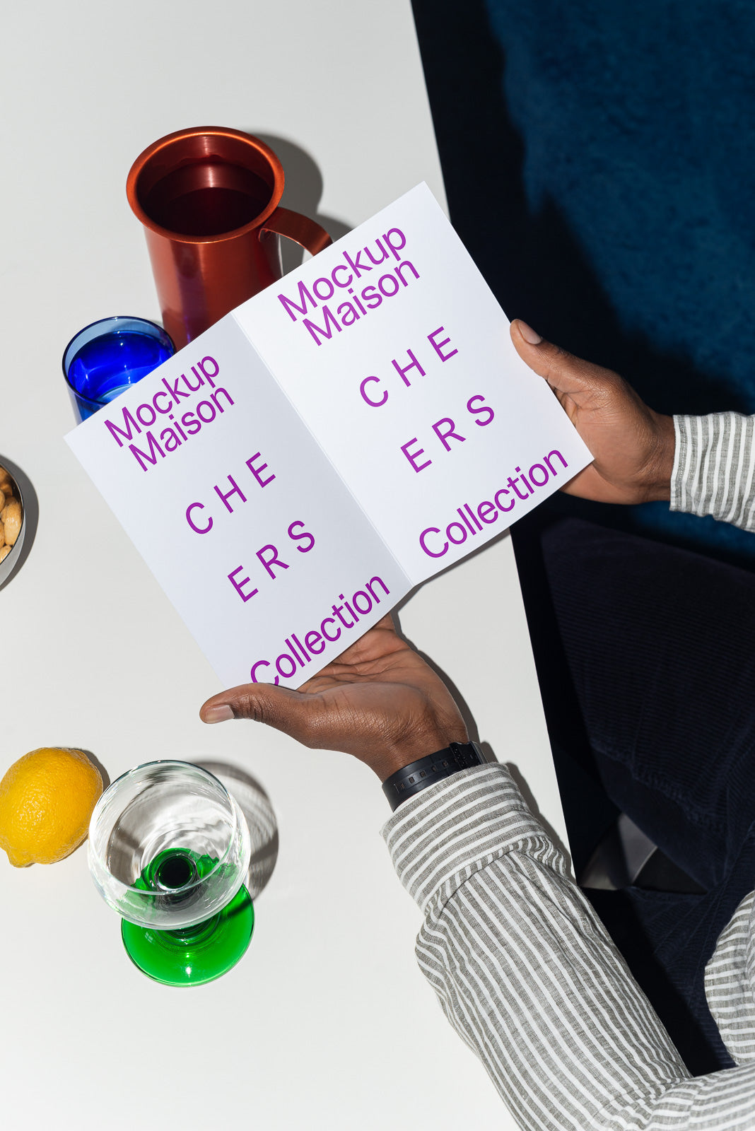 PSD mockup of a person holding an open brochure with purple text "Mockup Maison CHEERS Collection," next to a lemon, glass, and red mug on a table.