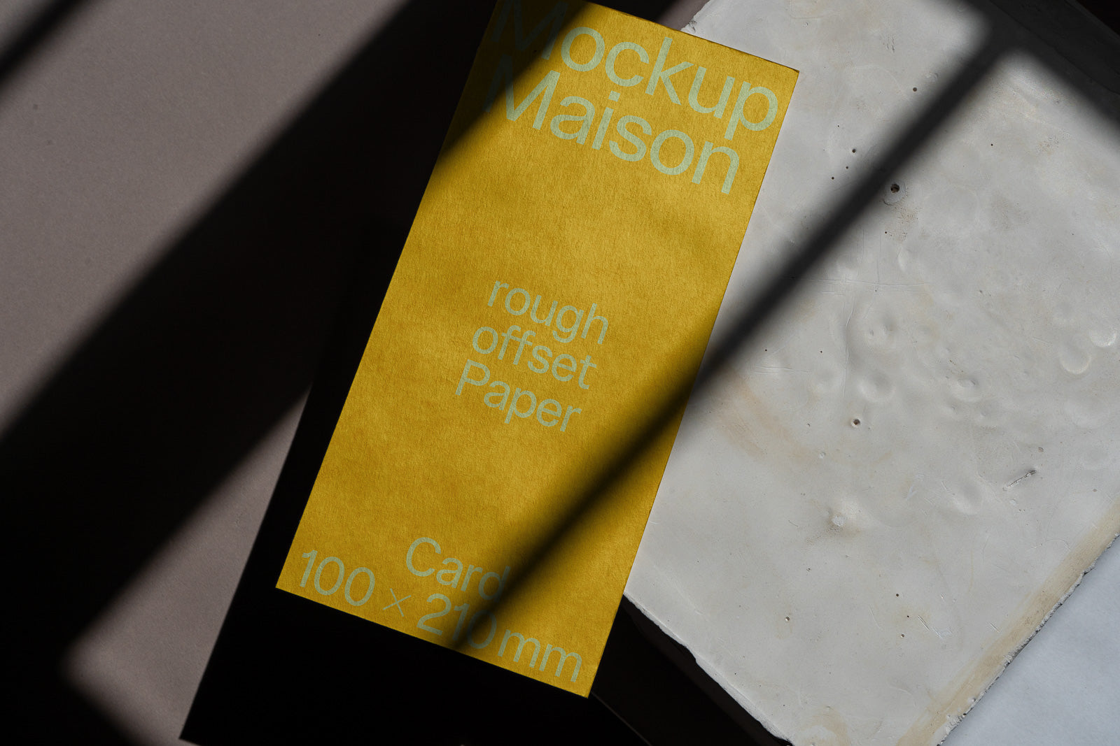 PSD mockup of a yellow card labeled "Mockup Maison" displaying text "rough offset Paper" and dimensions "100 x 210 mm," placed on a textured surface with shadow play.