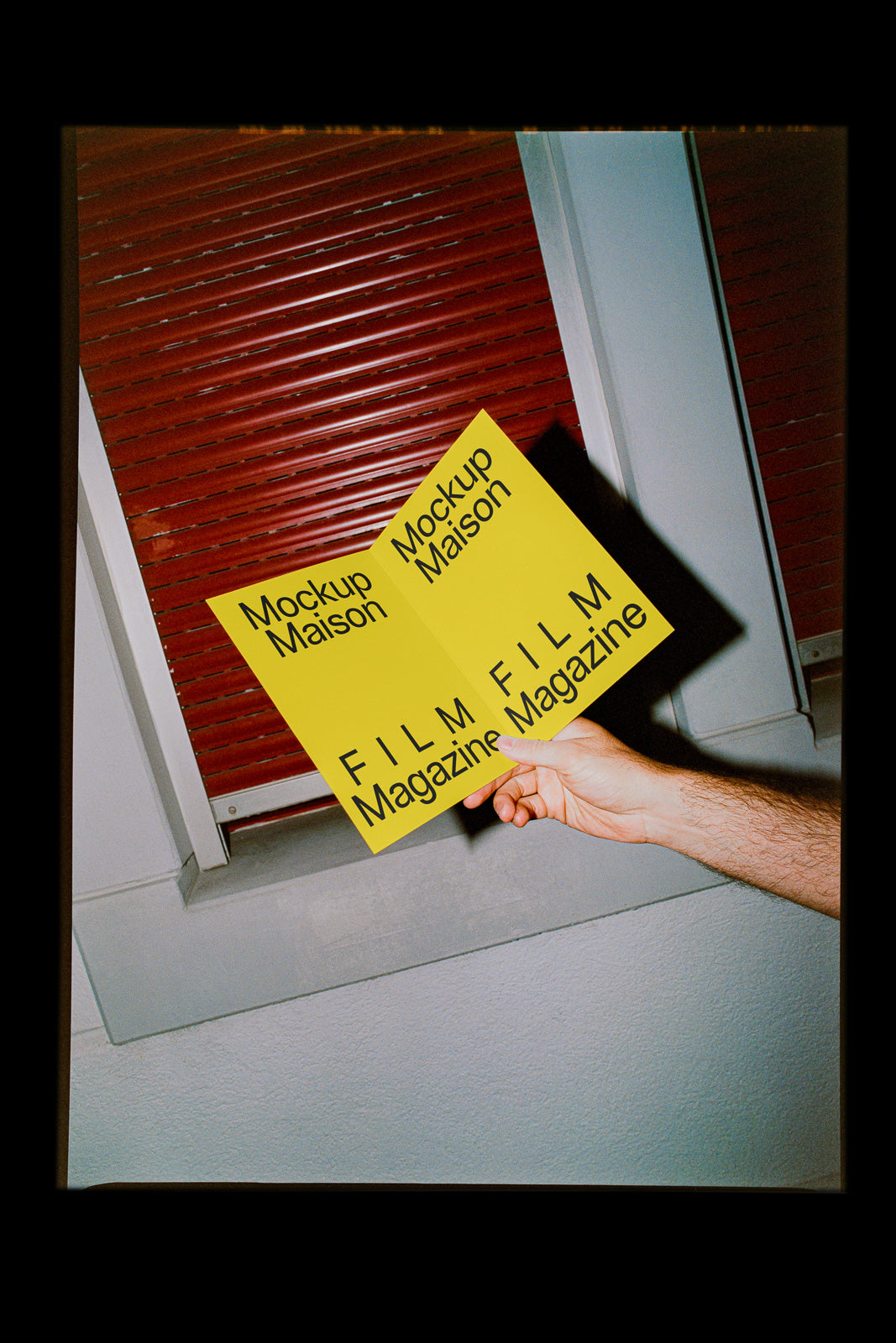 PSD mockup of a hand holding an open yellow booklet reading "Mockup Maison" and "FILM Magazine" against a red shutter background.