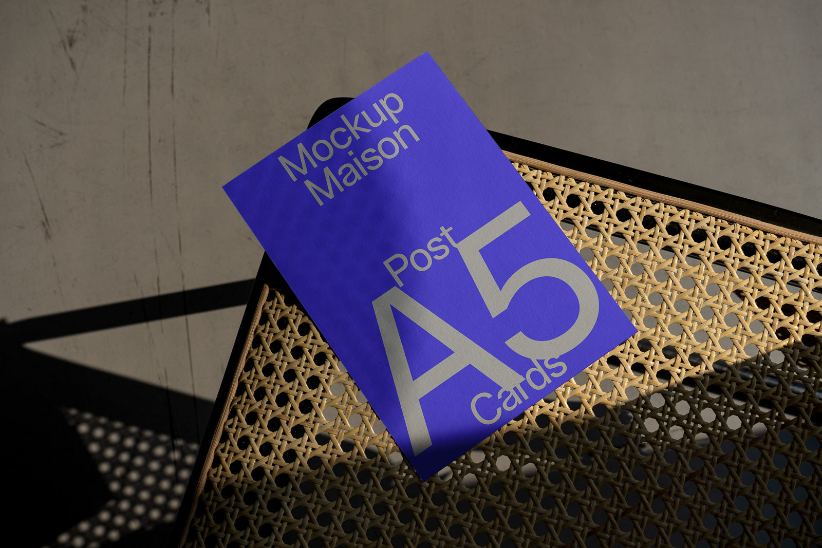 PSD mockup of an A5 postcard on a woven chair with sunlight casting shadows, featuring bold white text on a blue background.