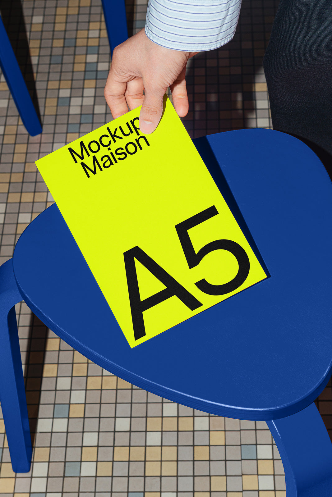 PSD mockup of a hand placing a bright yellow A5 document titled "Mockup Maison" on a blue triangular table with a tiled floor background.