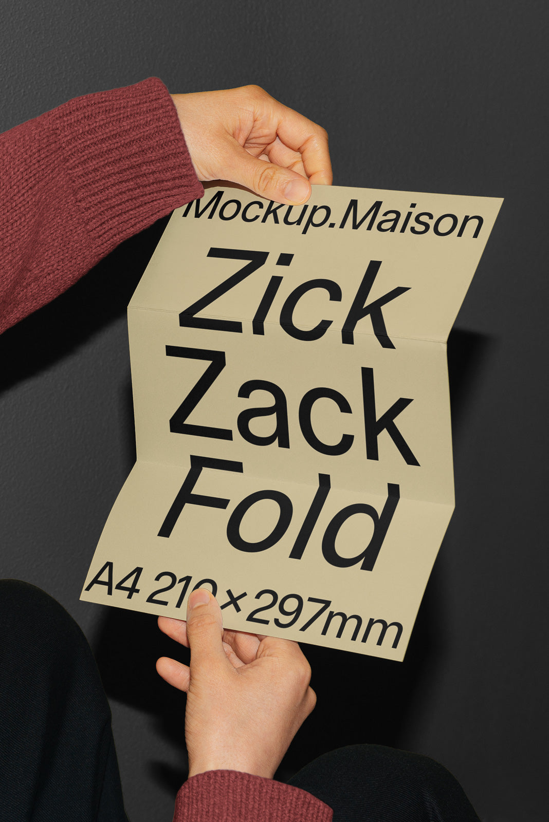 PSD mockup of a person holding a beige paper with bold black text "Mockup.Maison Zick Zack Fold A4 210x297mm" against a dark background.