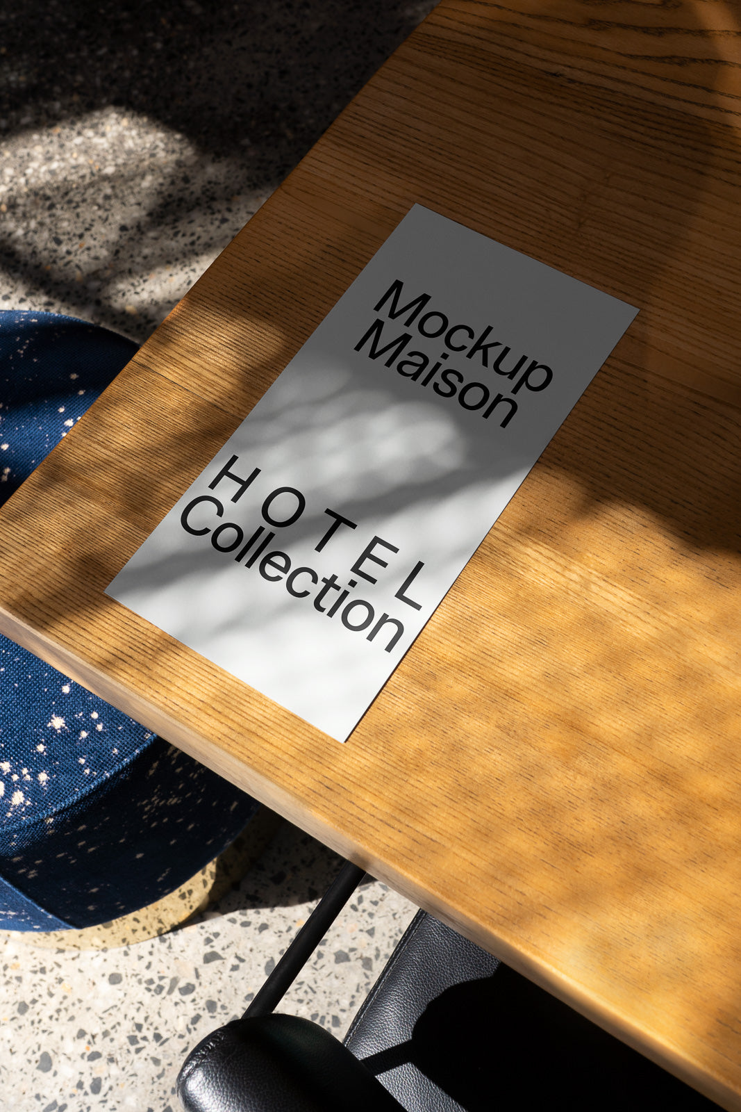 PSD mockup of a printed card with "Mockup Maison" and "HOTEL Collection" placed on a sunlit wooden table next to a blue speckled stool.