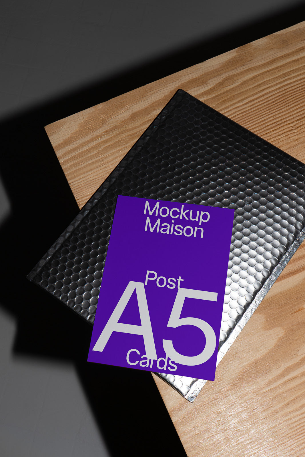 PSD mockup of a purple A5 card labeled "Mockup Maison" resting on a black bubble mailer on a wooden surface.