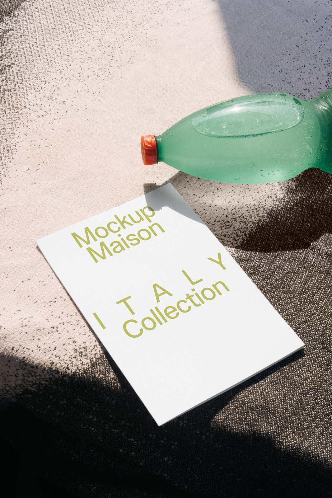 PSD mockup of a white card featuring "Mockup Maison Italy Collection" text in green, next to a green bottle with red cap, on a textured surface.