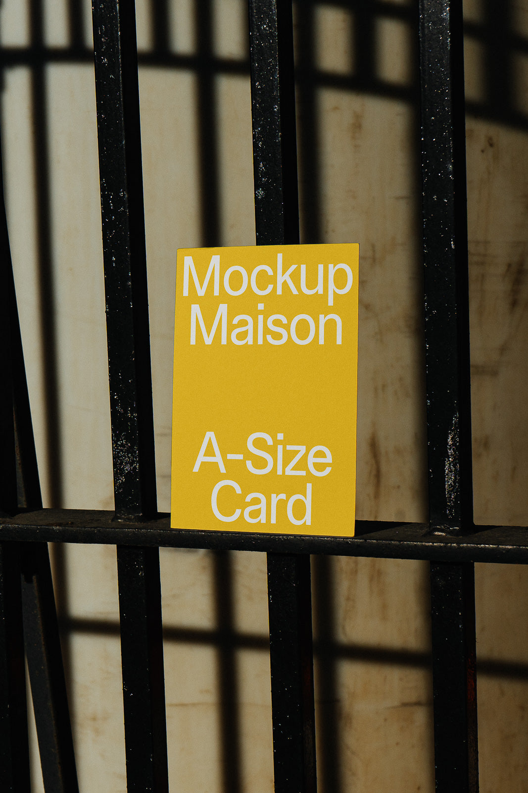 PSD mockup of a yellow A-size card labeled "Mockup Maison," positioned against a vertical metal railing with shadows cast on a textured background.