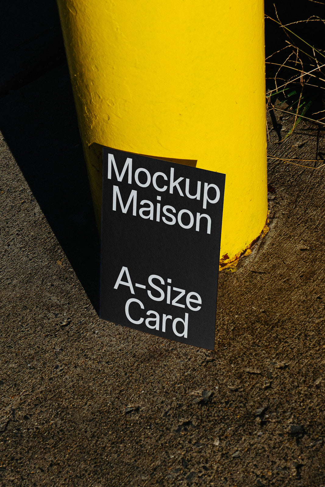 PSD mockup of an A-size card placed on the ground, leaning against a bright yellow cylindrical object, casting a shadow on the pavement.