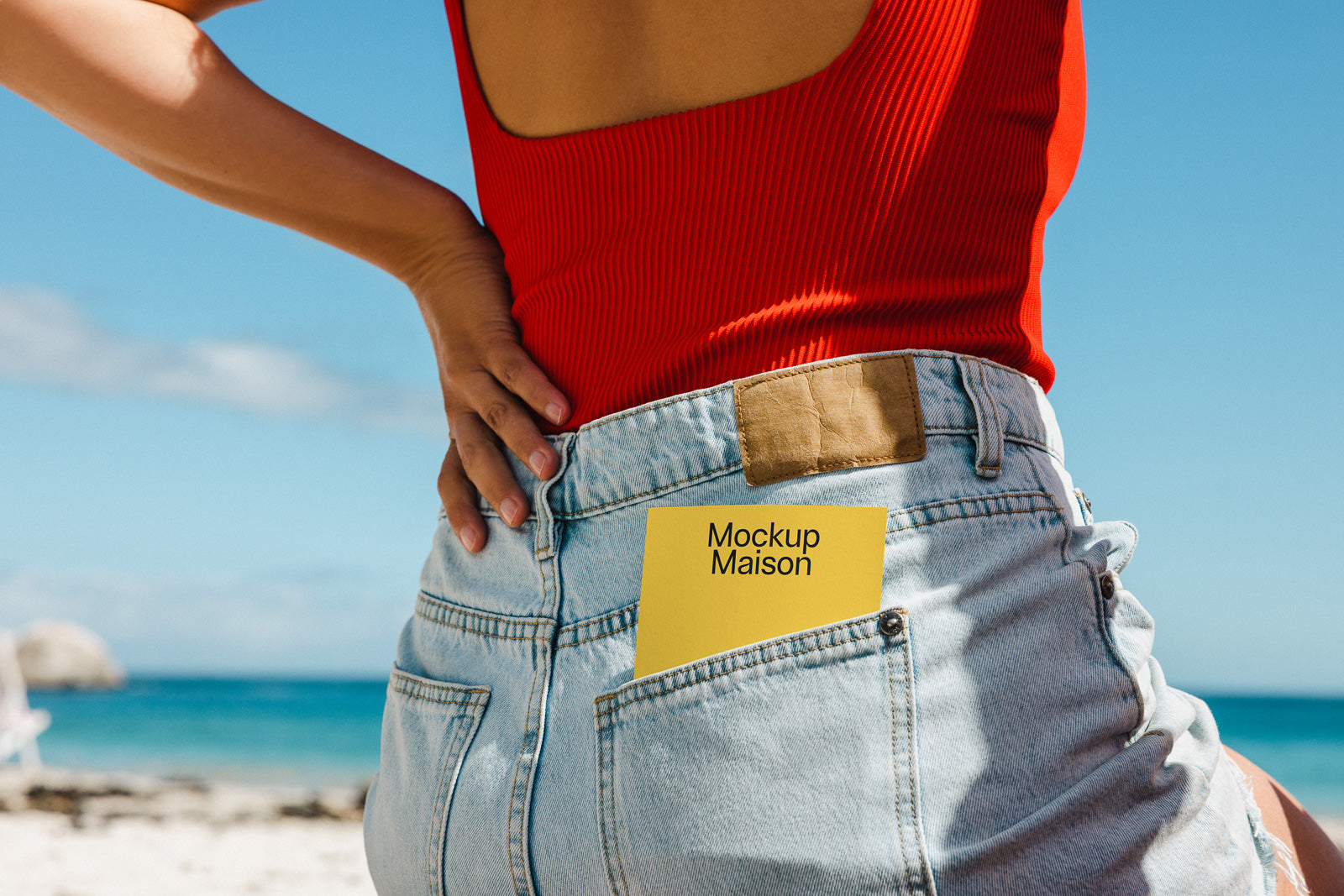 PSD mockup of a yellow card labeled "Mockup Maison" tucked into the back pocket of light blue denim jeans, worn with a red top, against a beach backdrop.