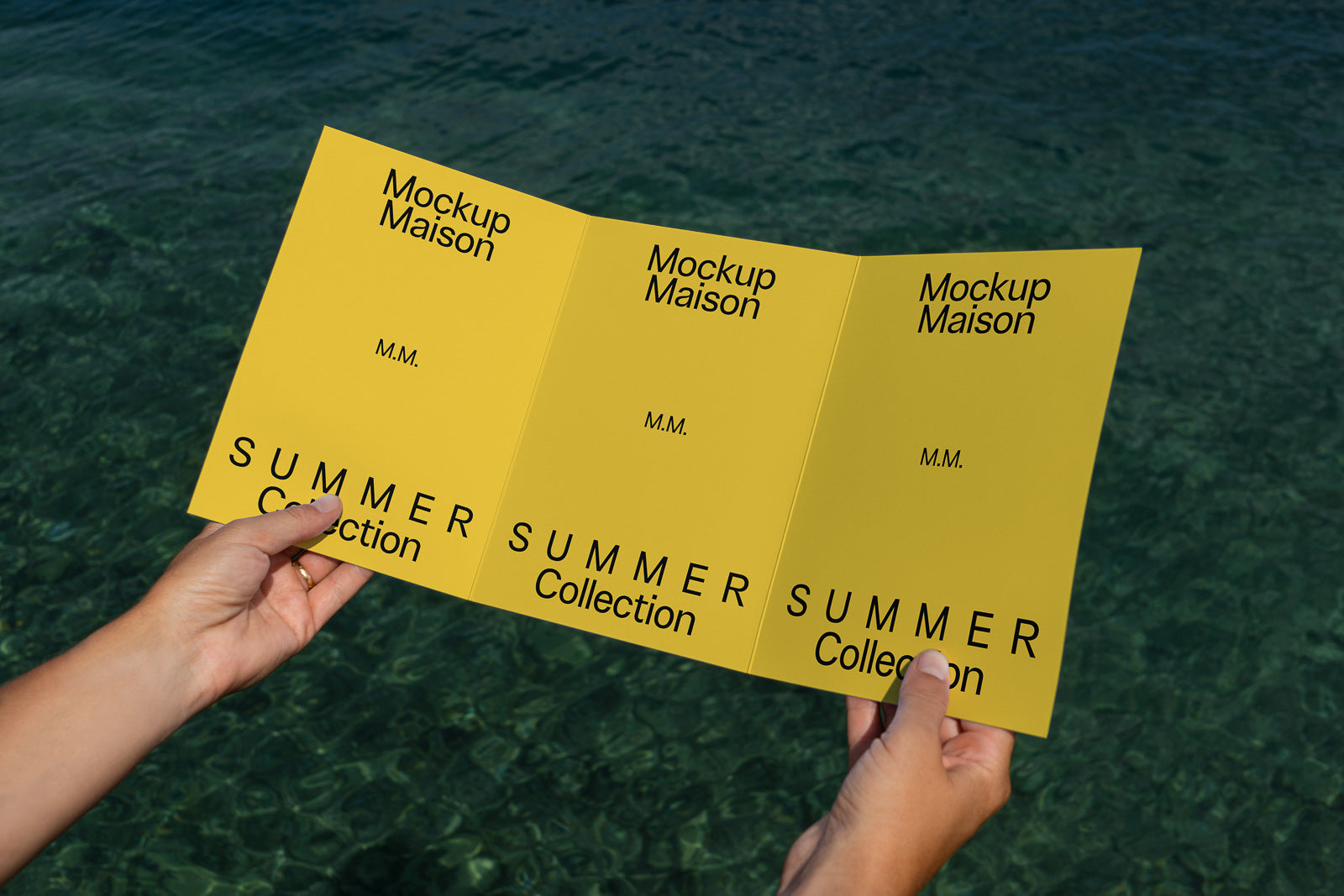 PSD mockup of a trifold brochure held by hands, displaying "Mockup Maison" and "Summer Collection" text on a yellow background over a clear water setting.