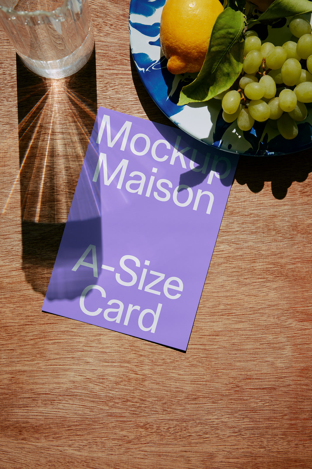 PSD mockup of an A-size card on a wooden table, with a glass casting light shadows and a plate holding a lemon and grapes.