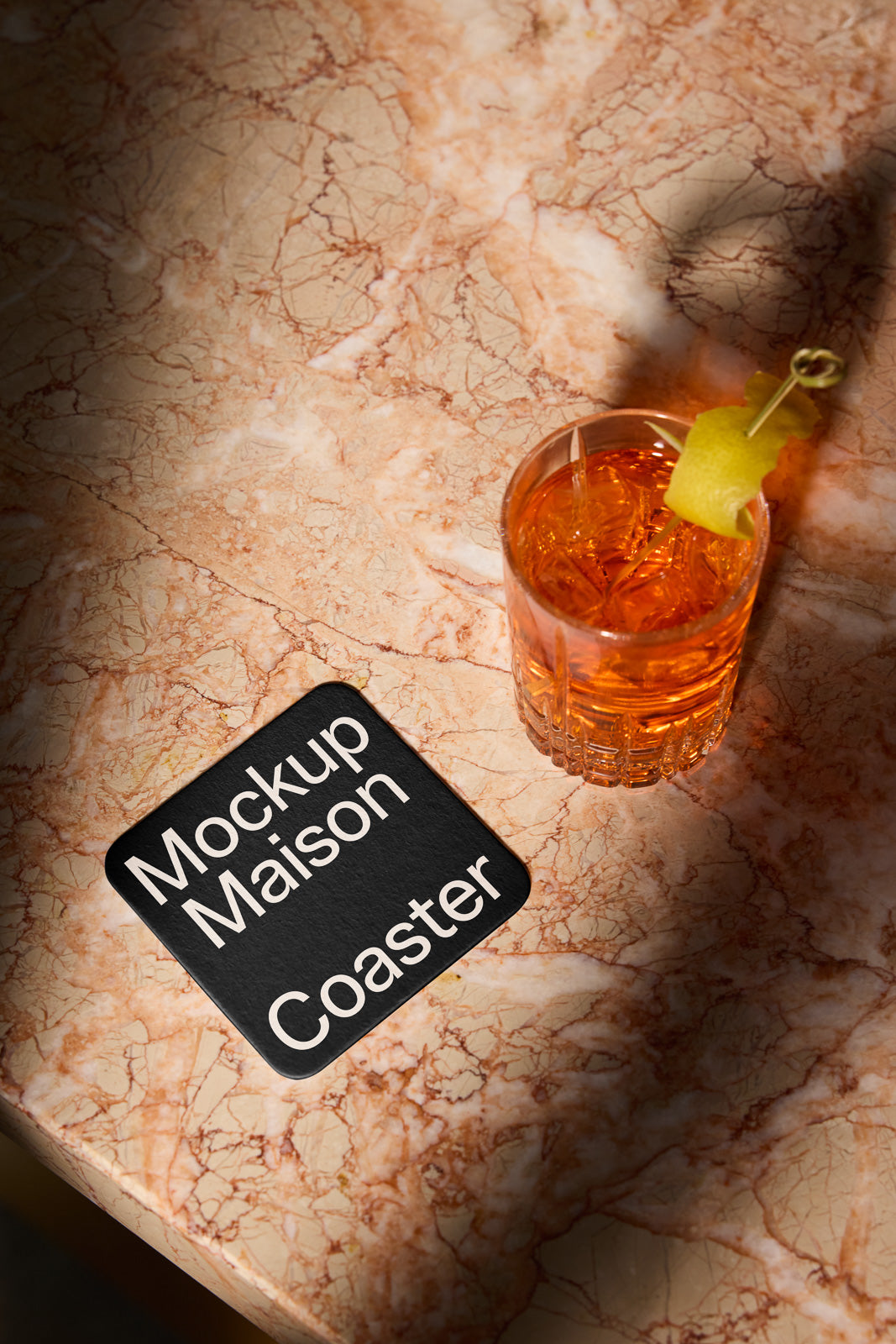 PSD mockup of a square coaster labeled "Mockup Maison Coaster" on a marbled surface next to a glass with an orange beverage and lemon garnish.
