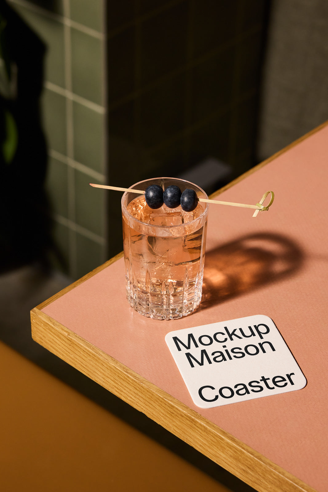 PSD mockup of a glass with ice and blueberries on a wooden skewer, beside a coaster with "Mockup Maison" text, placed on a pink tabletop.