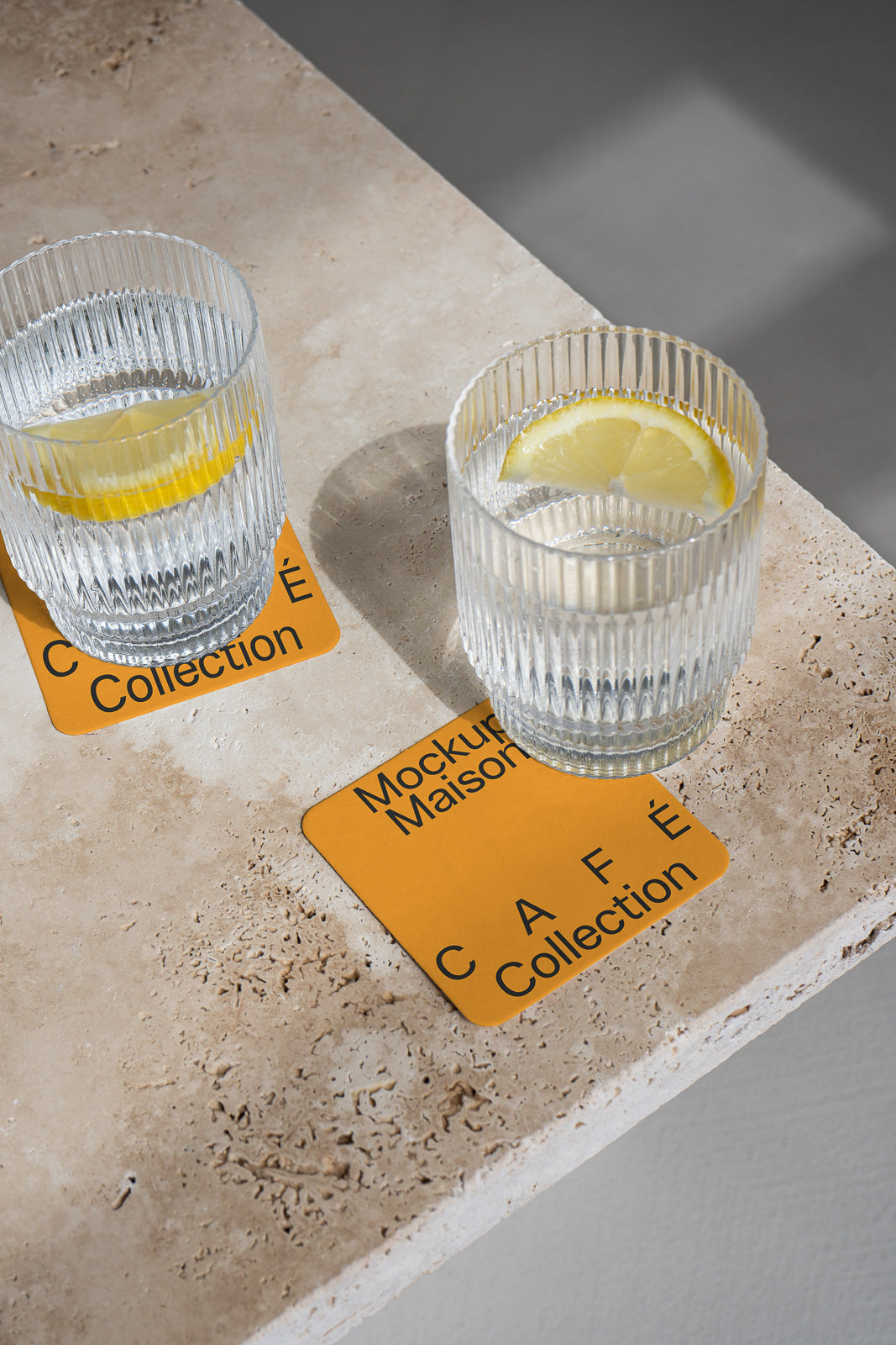 PSD mockup of two ribbed glasses with lemon slices on yellow coasters labeled "Café Collection" on a textured stone surface.