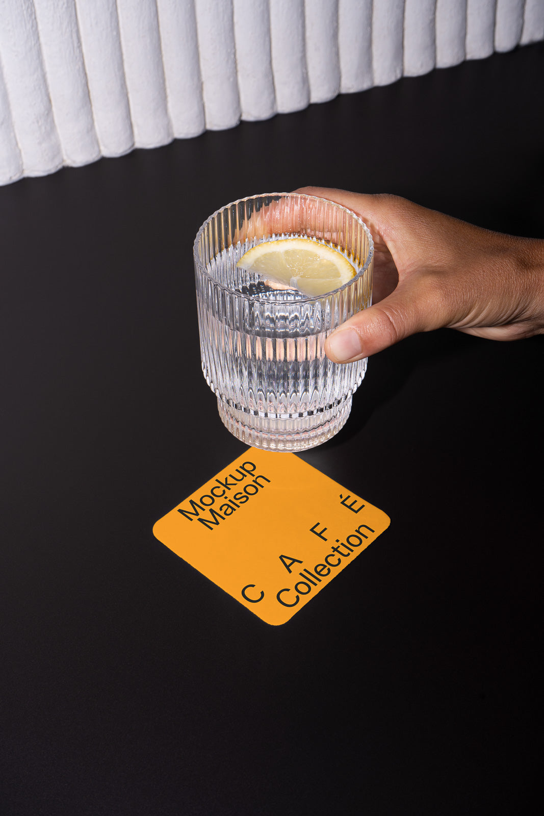 PSD mockup of a hand holding a glass with a lemon slice above a yellow square card labeled "Mockup Maison CAFE Collection" on a dark surface.